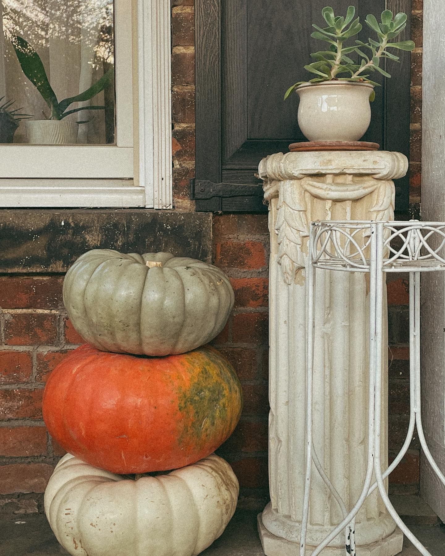 Stack of pumpkins next to a pillar with a potted plant on top and a window in the background.
