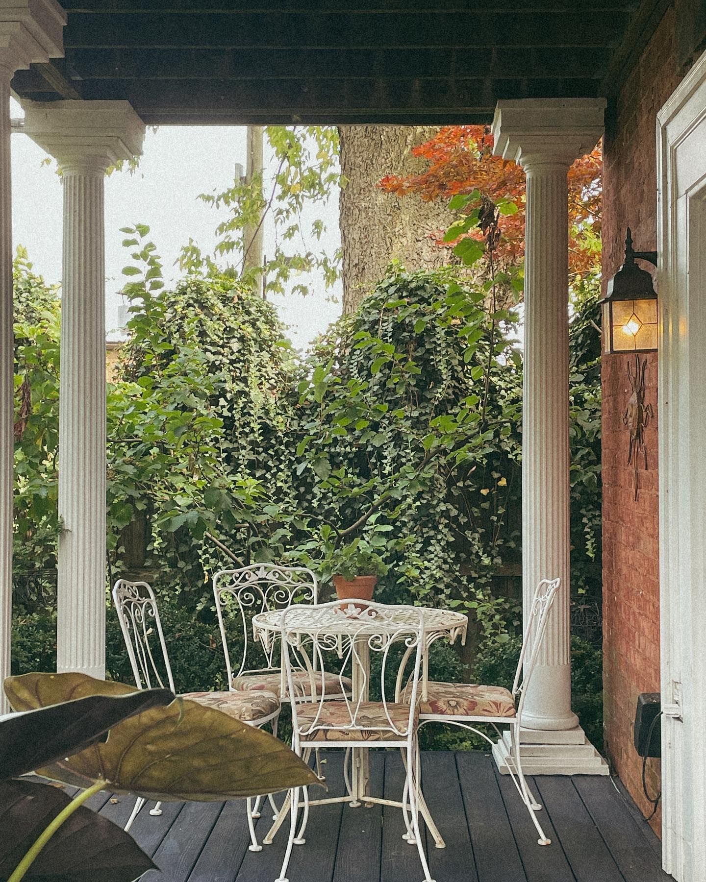 White table and chairs on a porch, framed by columns, lush greenery in background.
