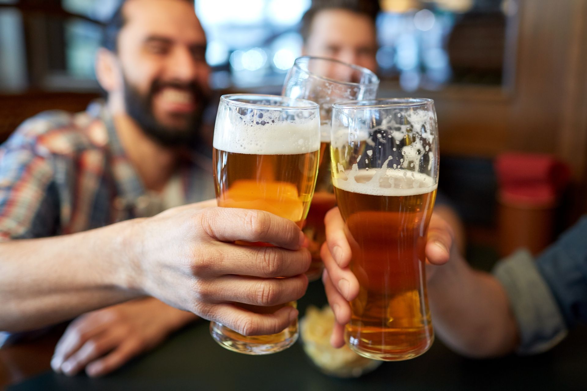 Three people clinking beer glasses in a pub, toasting. Beers are golden; a man smiles in the background.