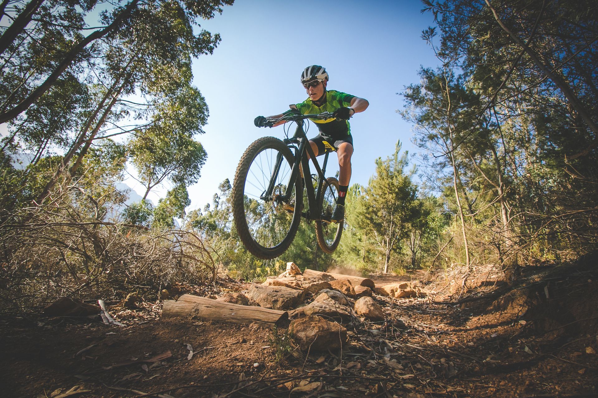 A mountain biker in green gear jumps over a rocky trail in a forest.