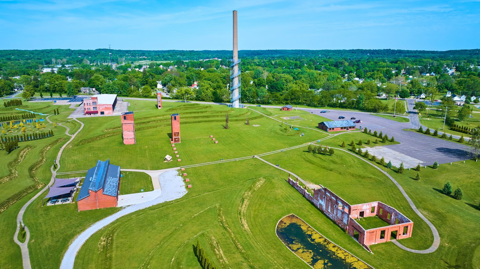 Aerial view of a green hill with red brick structures, a tall tower, and parking lot, set against a tree-lined horizon.