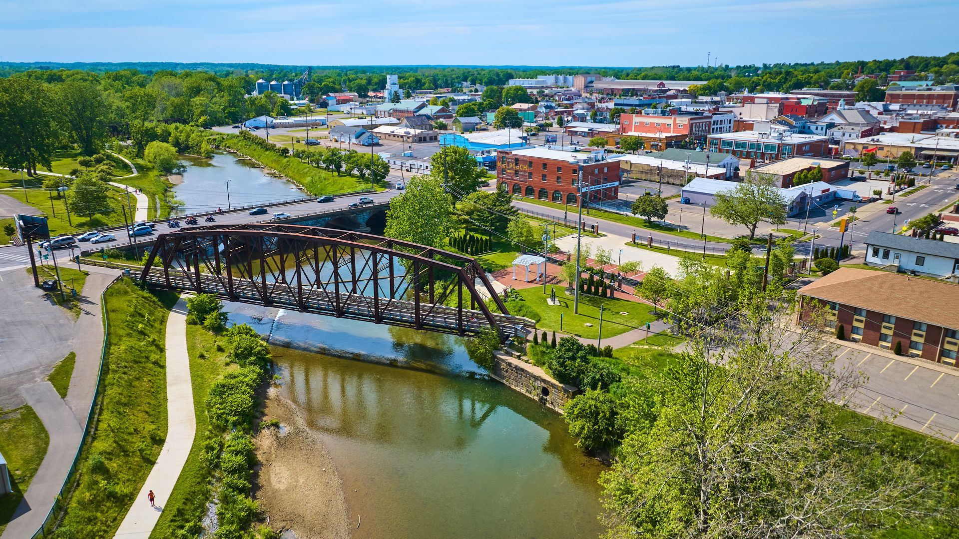 Overhead view of a town with a river, bridge, green trees, and buildings on a sunny day.