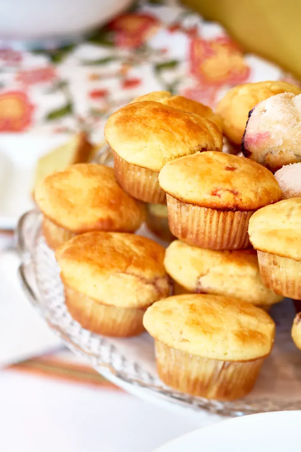 Pile of golden muffins on a glass cake stand, with a floral patterned tablecloth in the background.