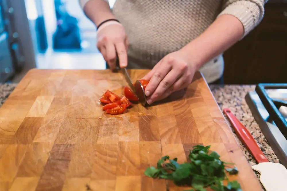 Person chopping a tomato on a wooden cutting board with a knife; cilantro is visible nearby.