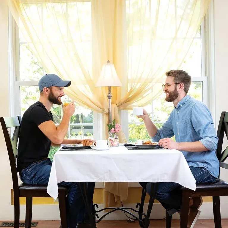 Two people smiling and having breakfast at a table near a window.