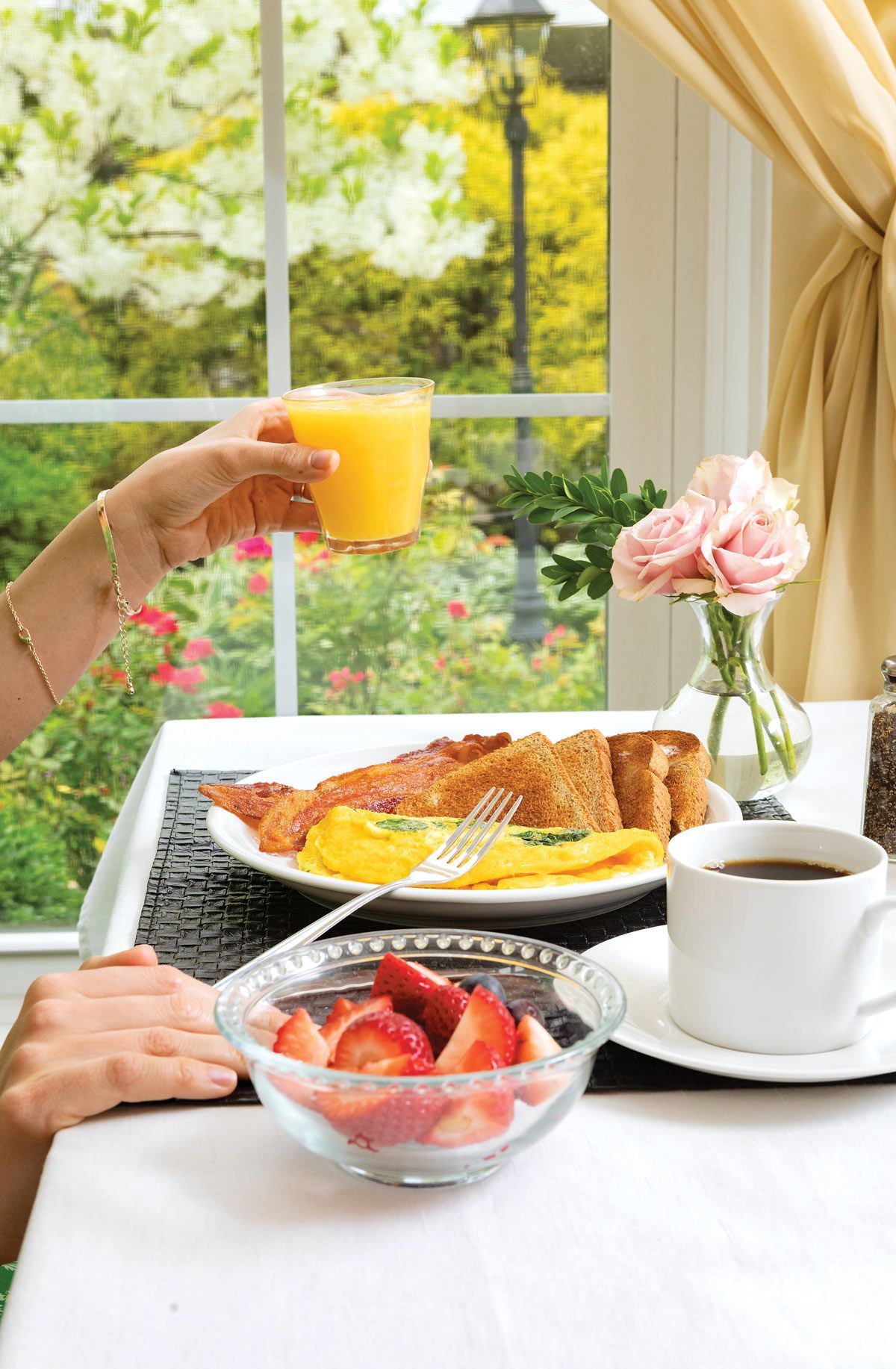Breakfast table with orange juice, bacon, eggs, toast, strawberries, and coffee next to a window with a garden view.