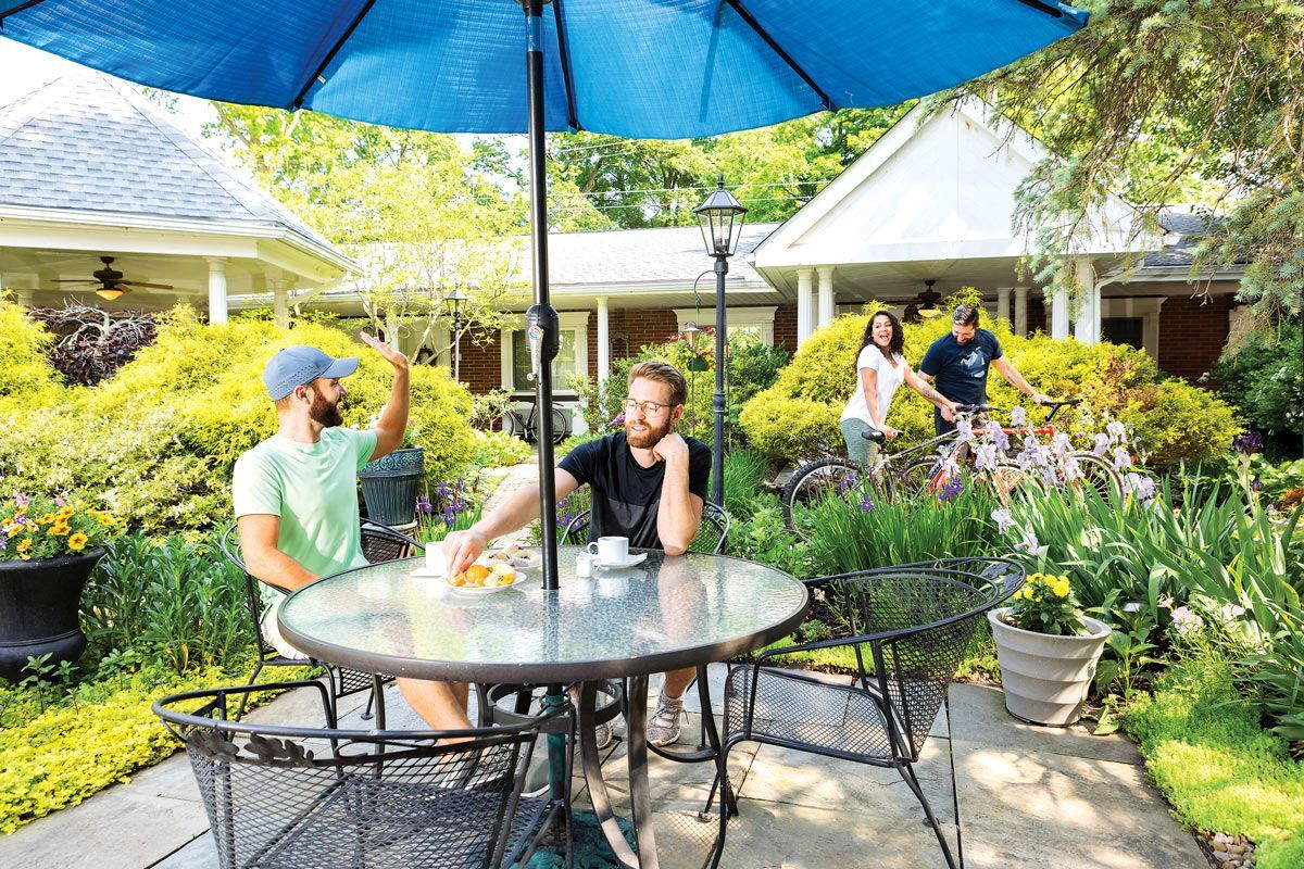 Two men at a cafe table under a blue umbrella in a garden, with people walking in the background.