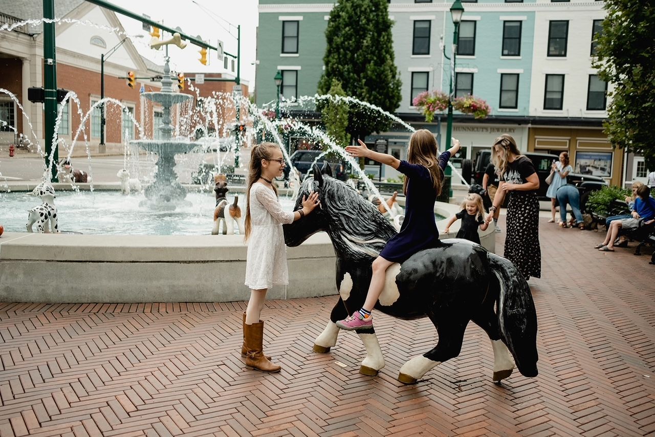 Children playing with a horse statue in front of a fountain in a town square.