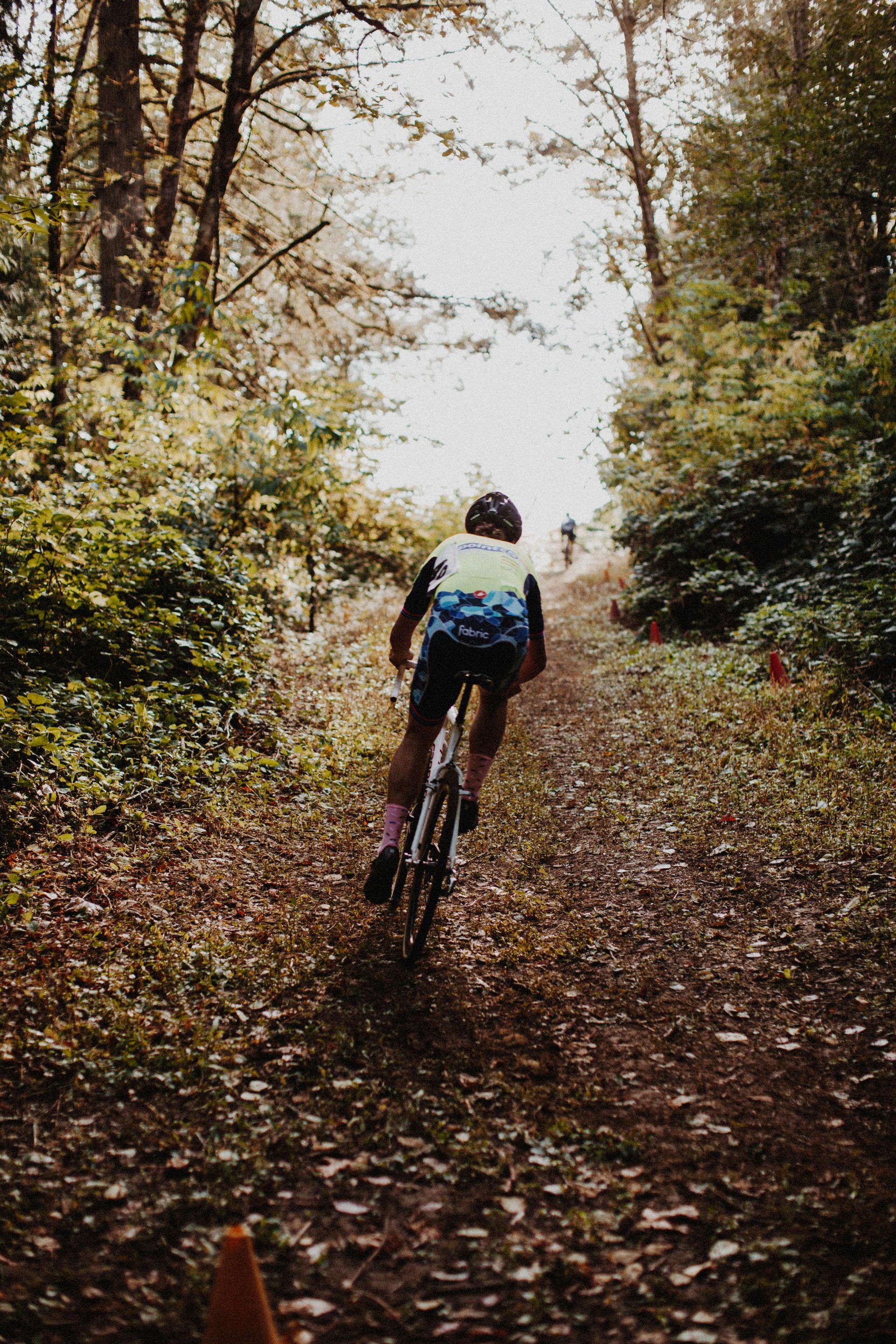 Cyclist riding a bicycle up a dirt path in a wooded area, with trees and foliage on either side.