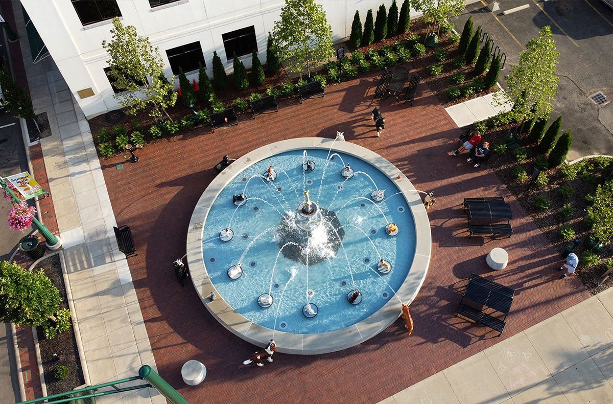 Overhead view of fountain in a brick plaza. People sit on benches around the fountain; some stand nearby.