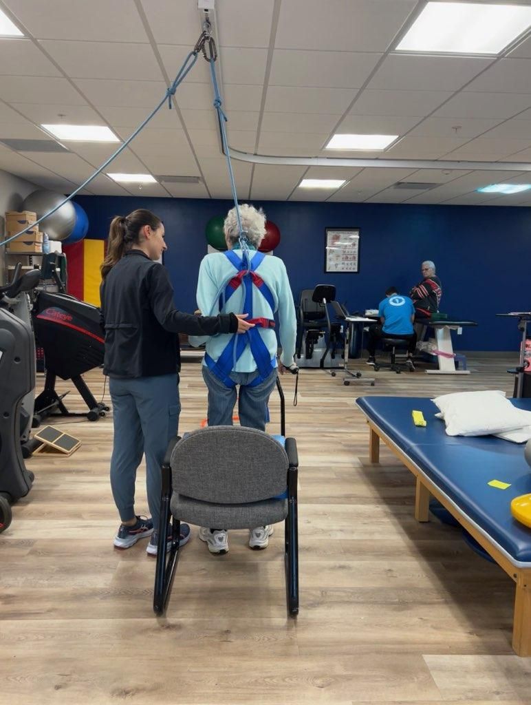 A woman is helping a woman wearing a safety harness walk in a gym.