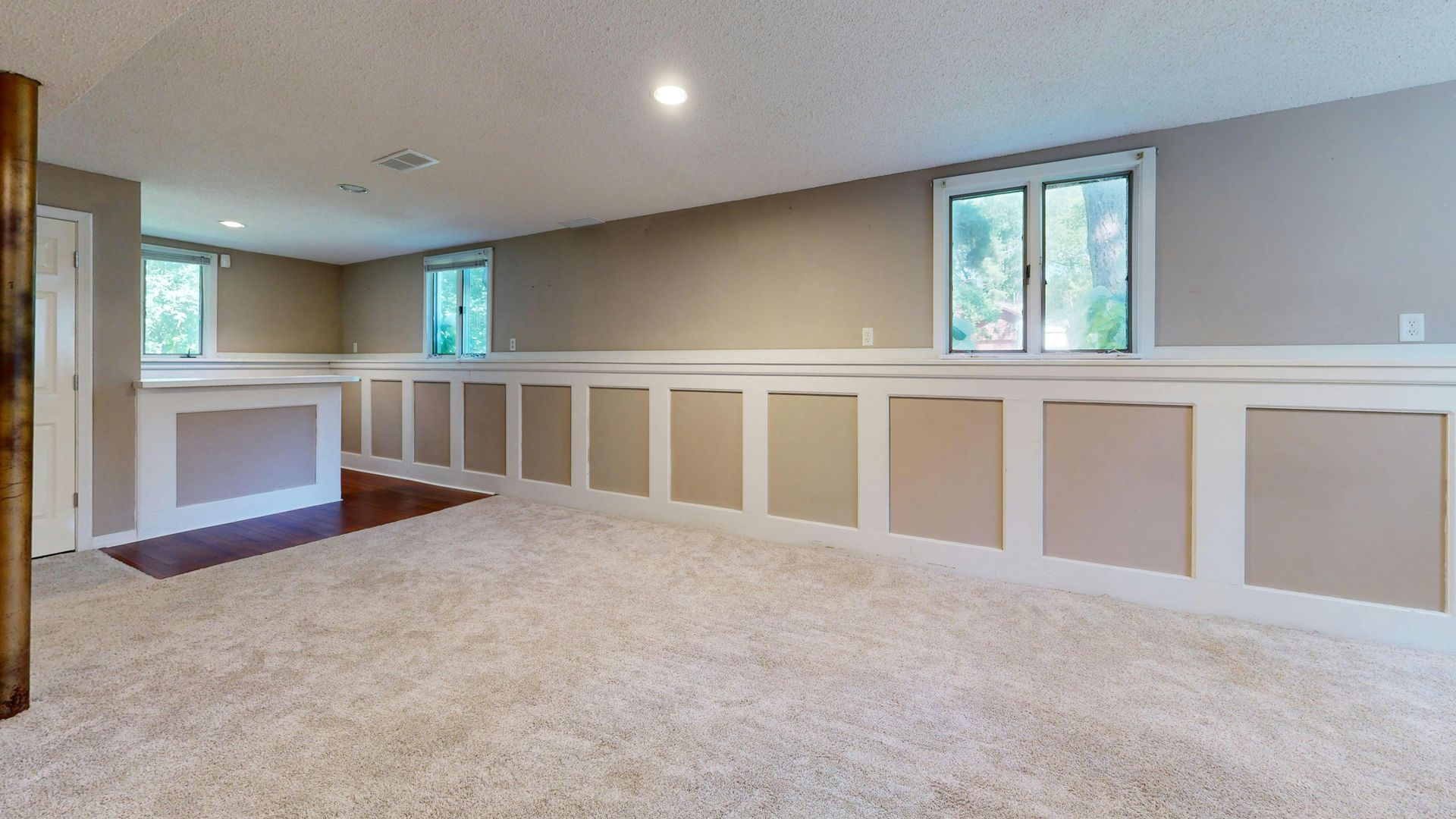 Bright carpeted empty room with beige walls, white wainscoting, and small windows.
