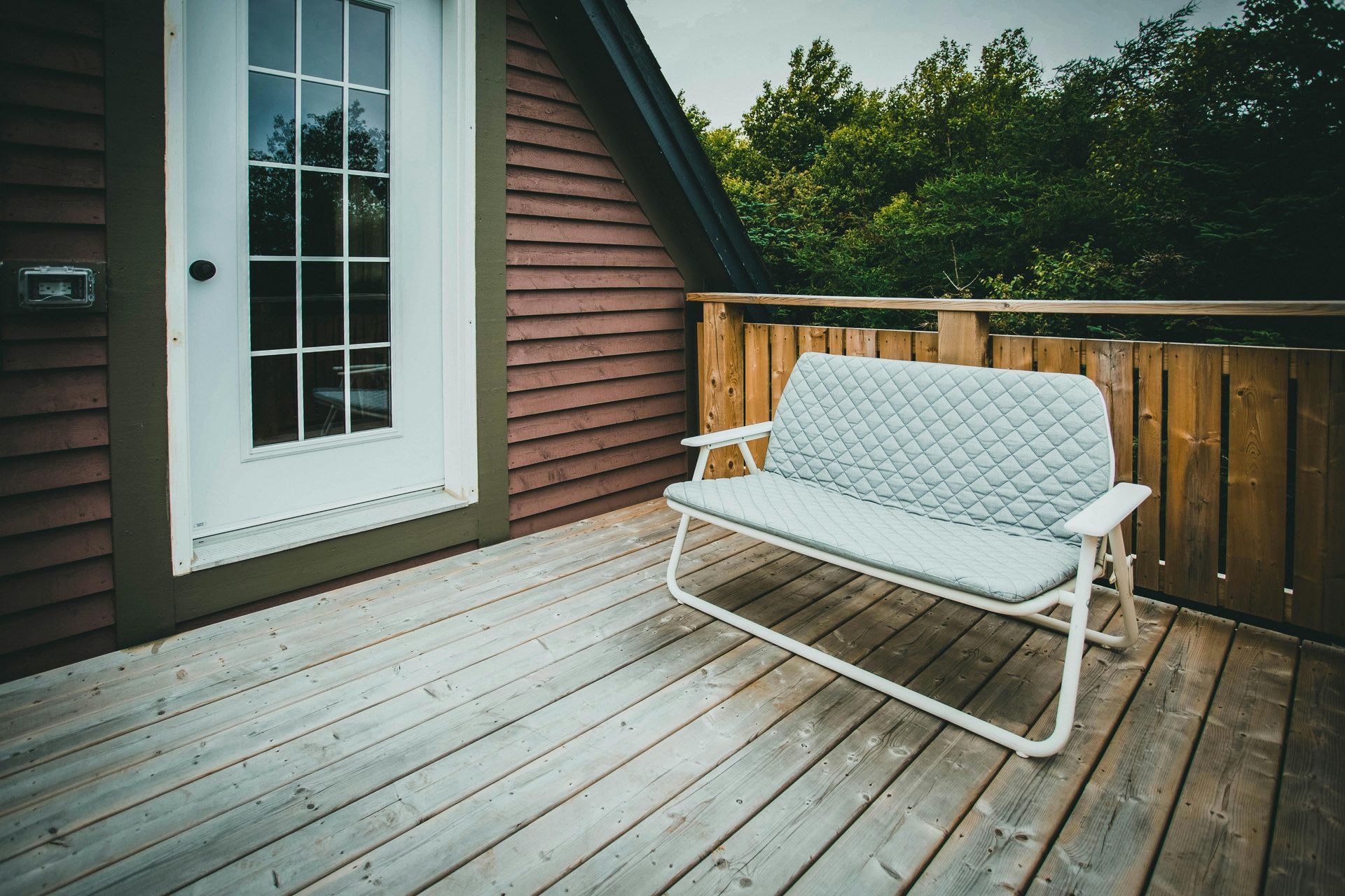 White bench on a wooden deck beside a house door and railing, with trees in the background