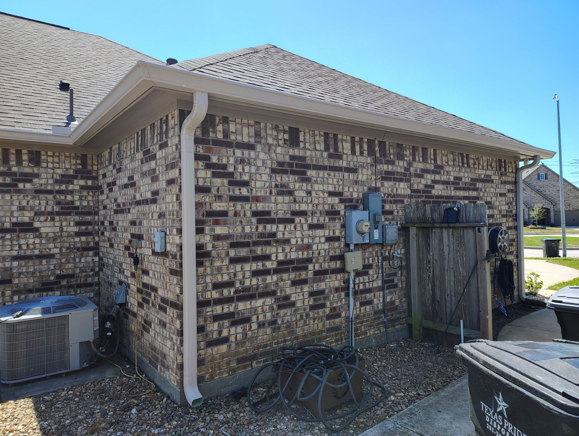Brick building exterior with tan trim, gutters, and a brown shingled roof, AC unit, and electrical boxes.