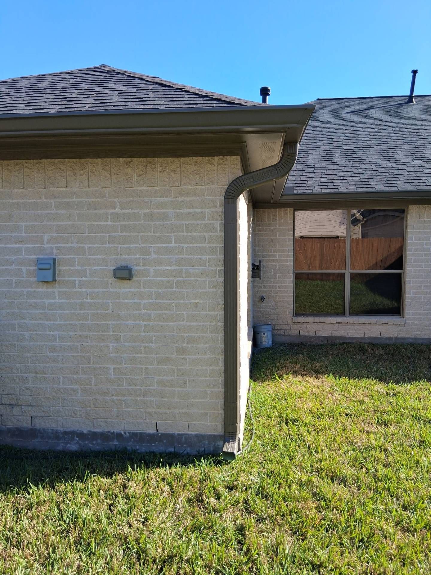 Beige brick house exterior with dark roof and green grass in sunlight.