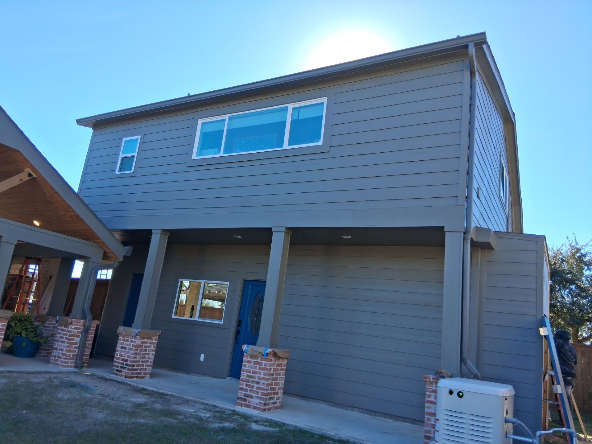 Two-story house with gray siding, blue door, and red brick columns. Bright sunny day.