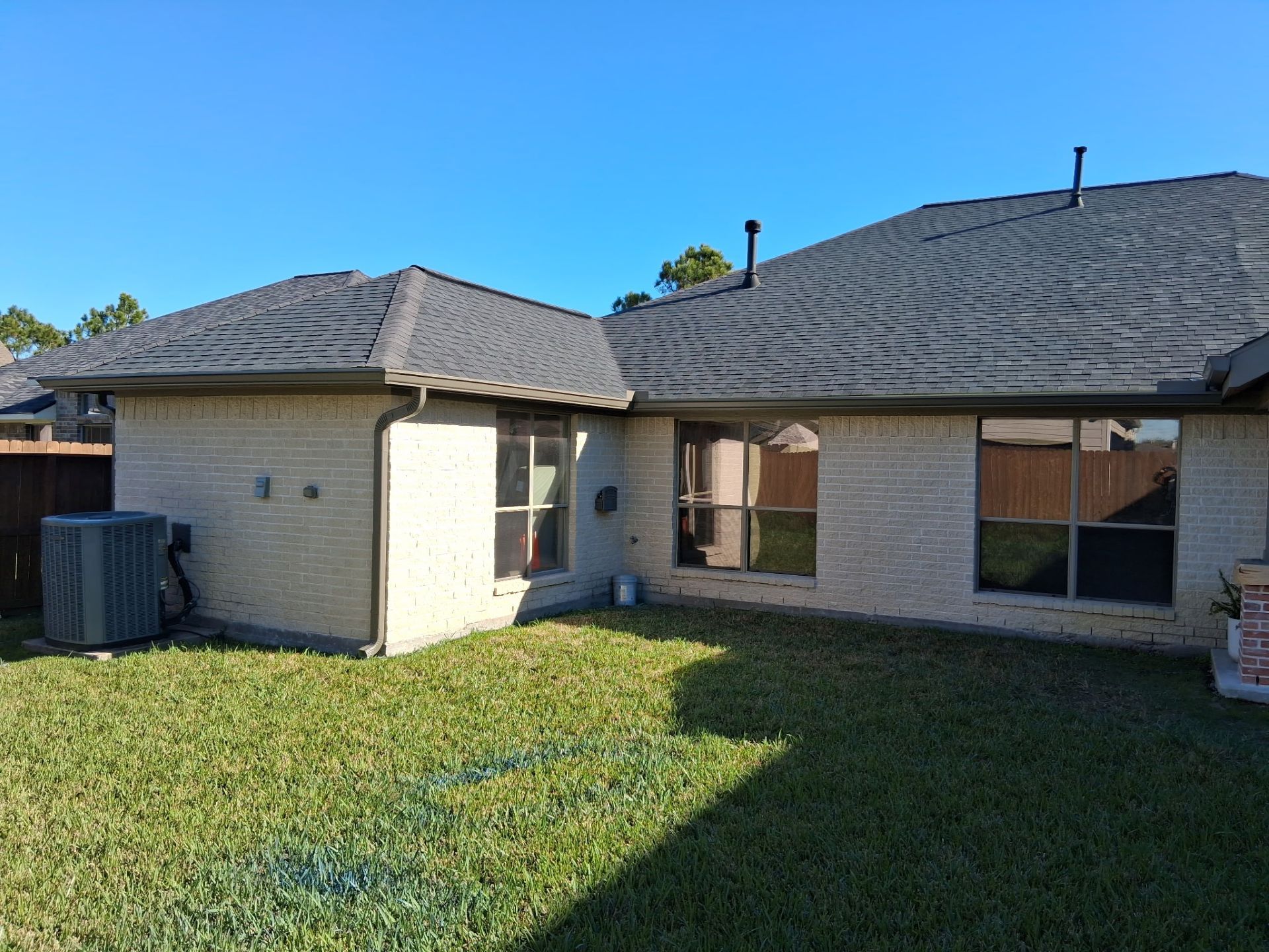 Backyard view of a house with light brick, windows, and a dark shingled roof under a clear blue sky.