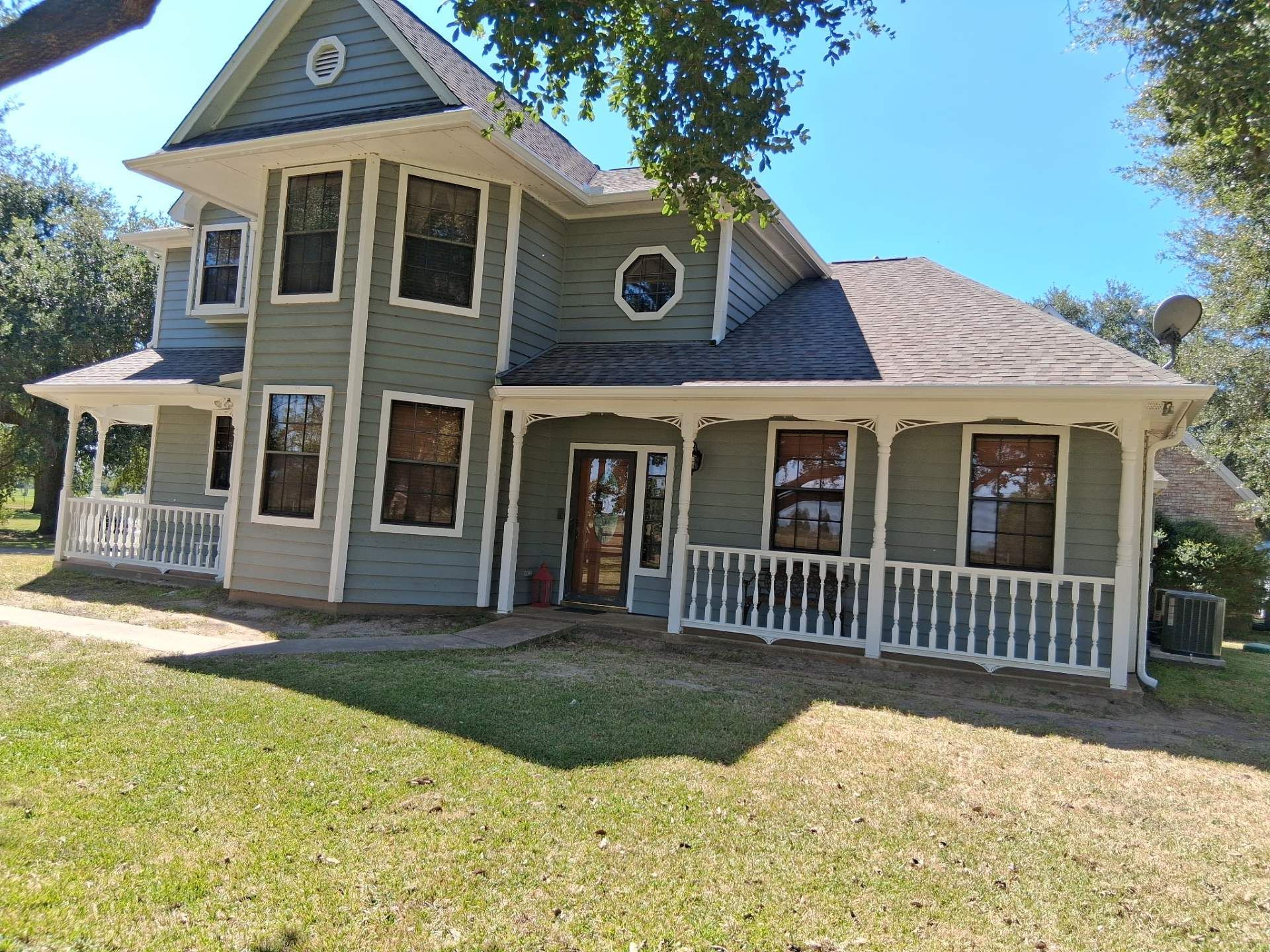 Two-story green house with white trim and a wrap-around porch on a sunny day.