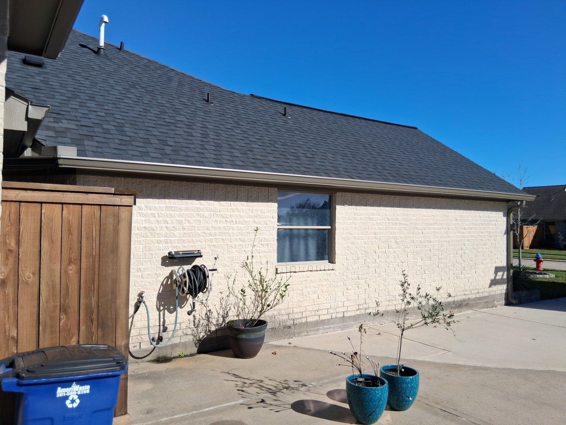 Brick building with dark roof, window, wooden fence, hose, potted plants, and blue sky.