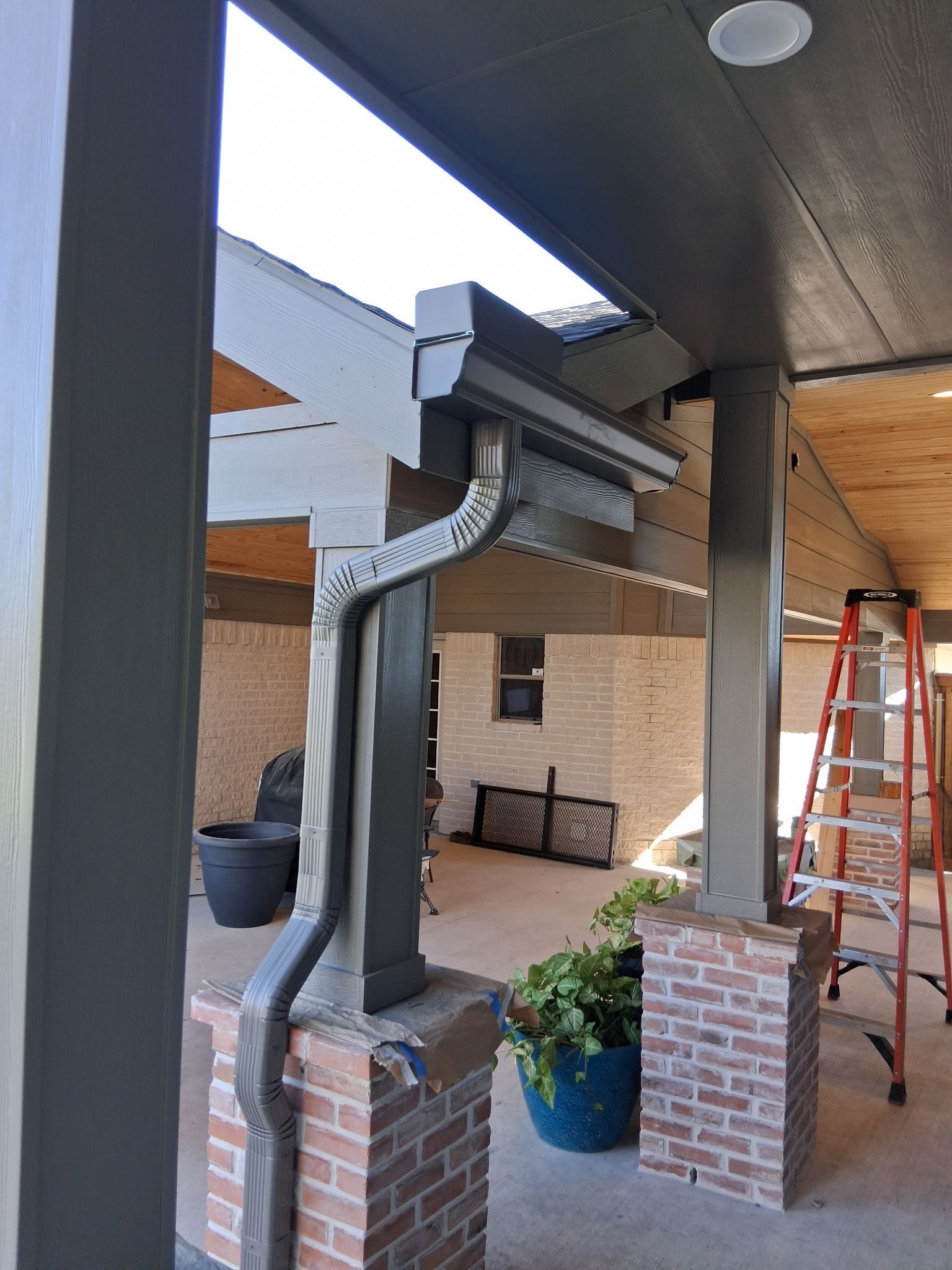 Exterior view of a porch with a decorative gutter. Gray pillars and accents, brick base, ladder visible.