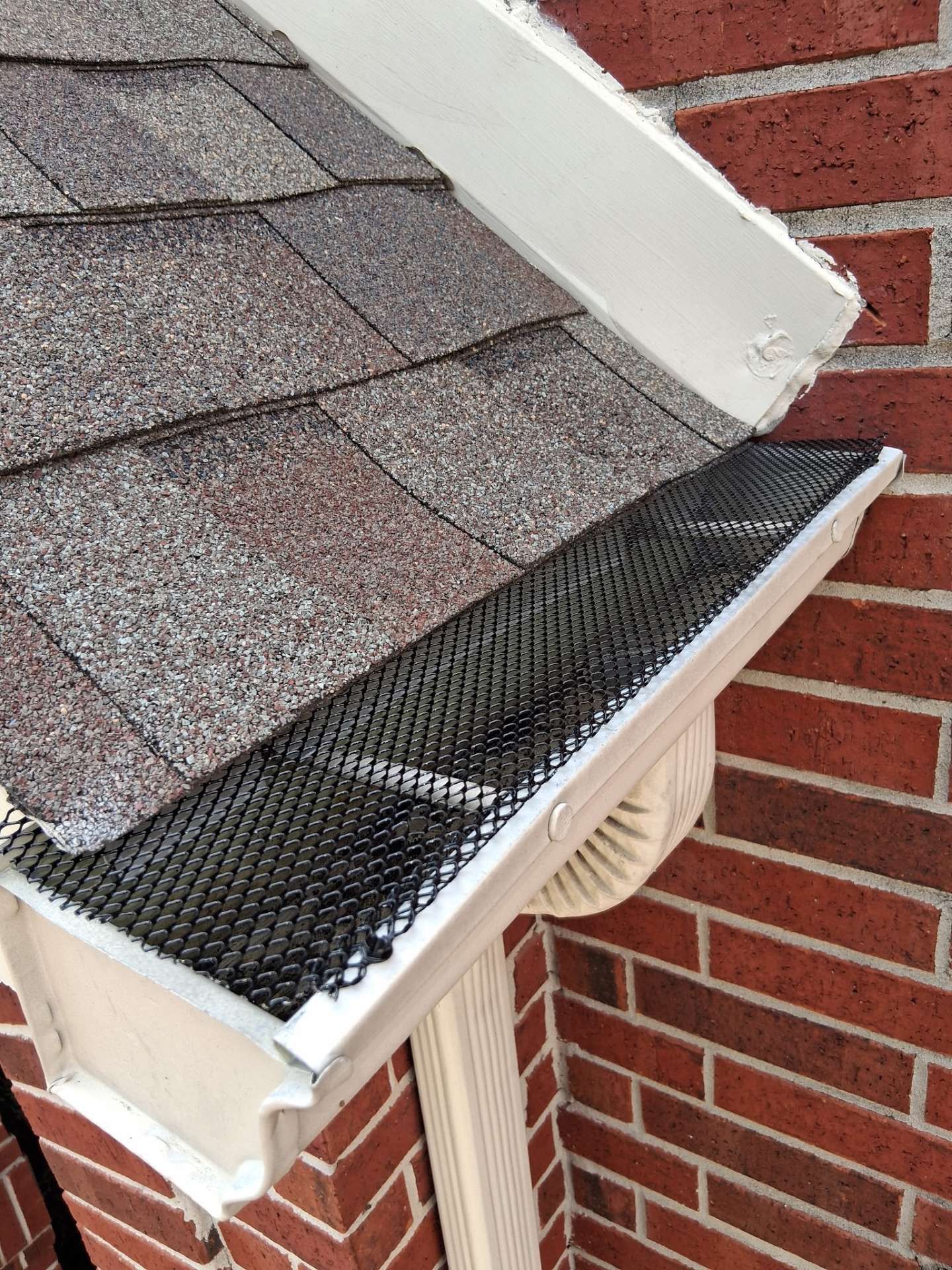 Black mesh gutter guard on a white gutter below brown shingles and a brick wall.