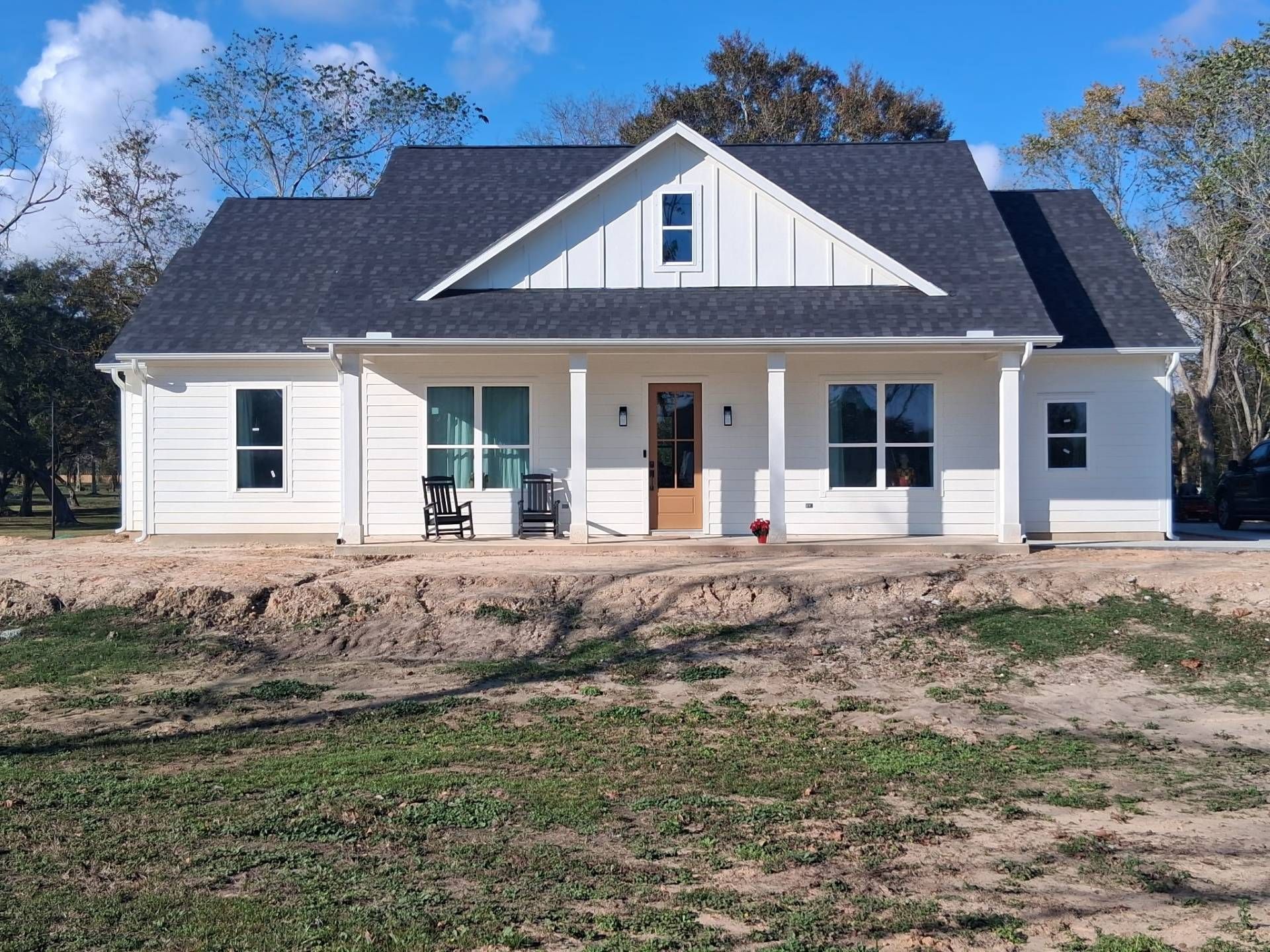 White farmhouse with black roof, brown door, and porch under a blue sky.