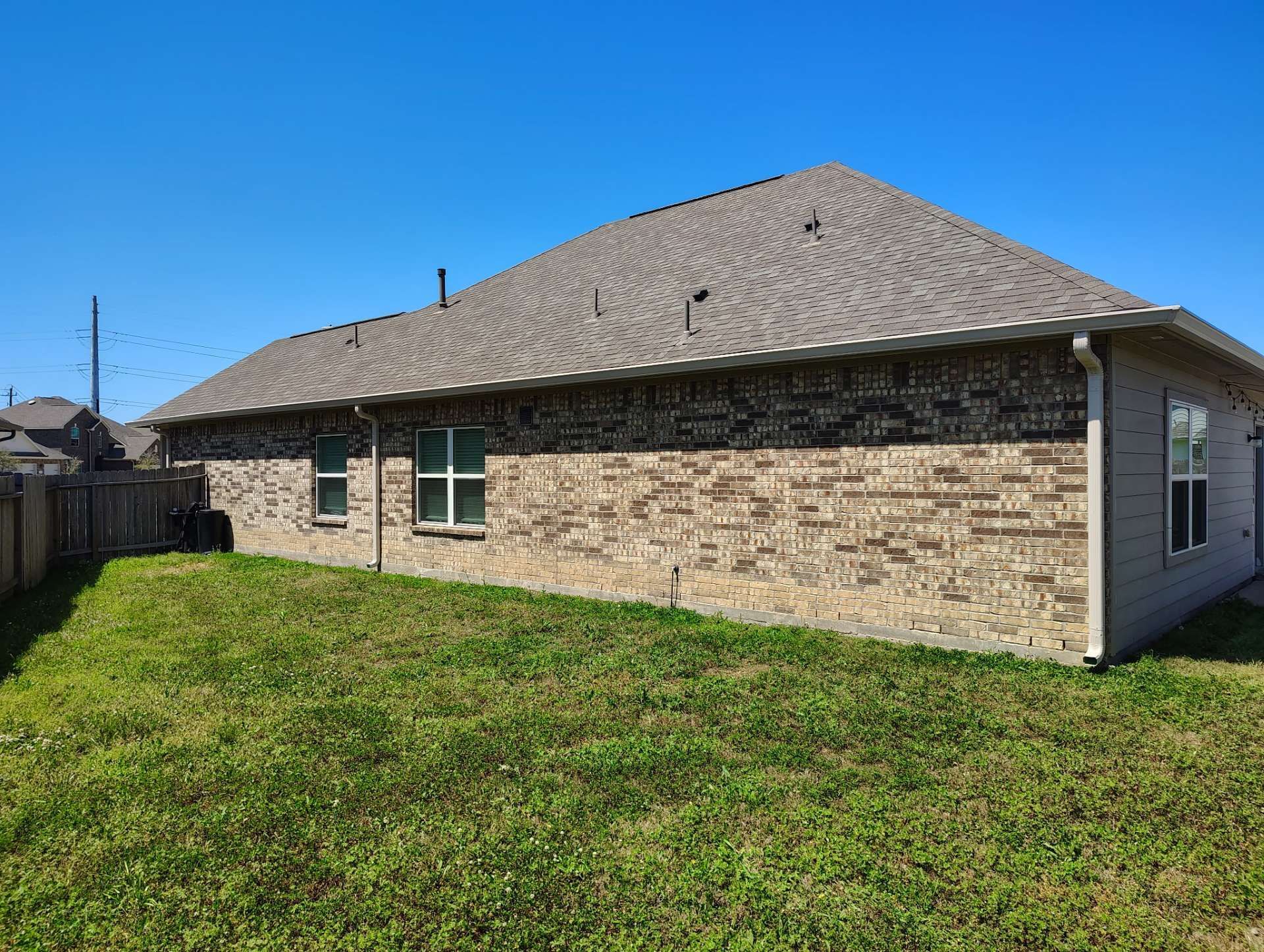 Brick house with brown roof, windows, and grassy backyard under a blue sky.