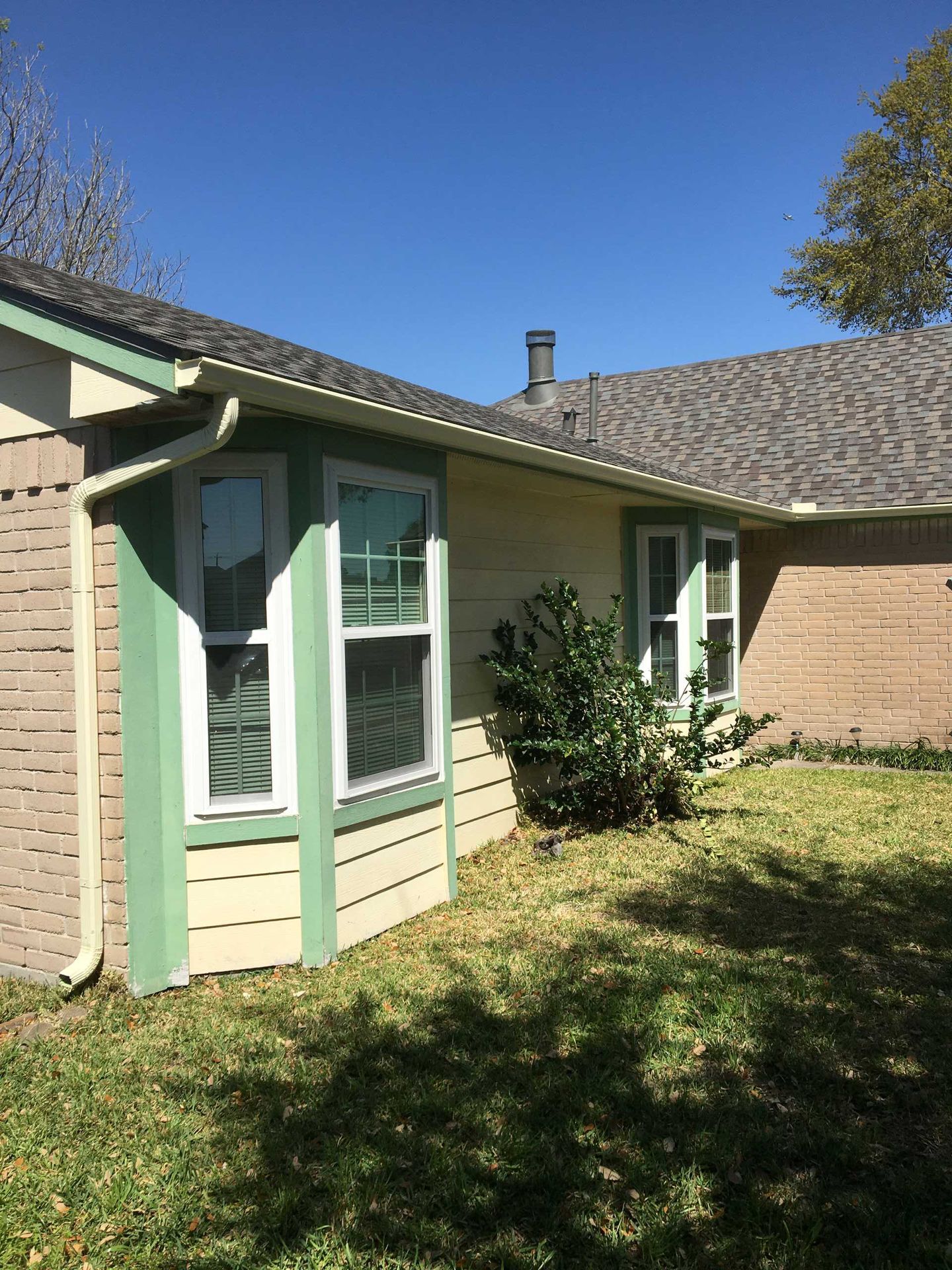 Exterior of a house with bay windows, beige siding, green trim, and a brick wall, under a clear blue sky.