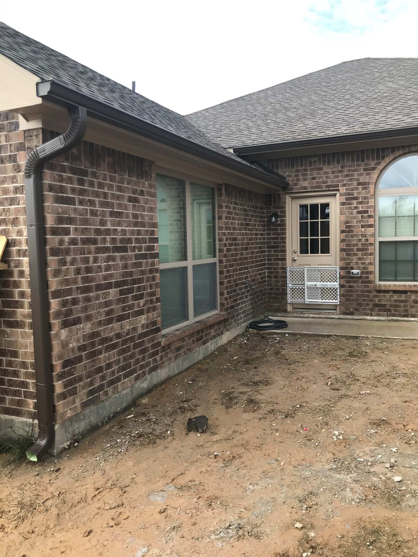 Brick house exterior with dark brown gutters, a window, and a door on a dirt lot.