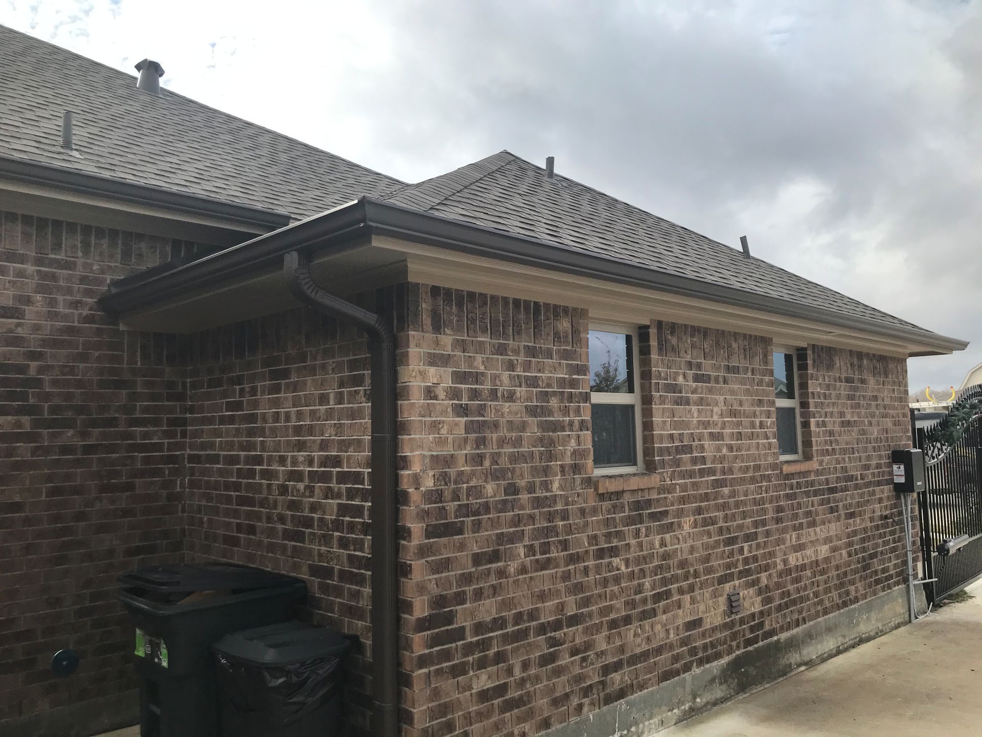 Brown brick house exterior with brown gutters and roof. Windows and trash cans visible.