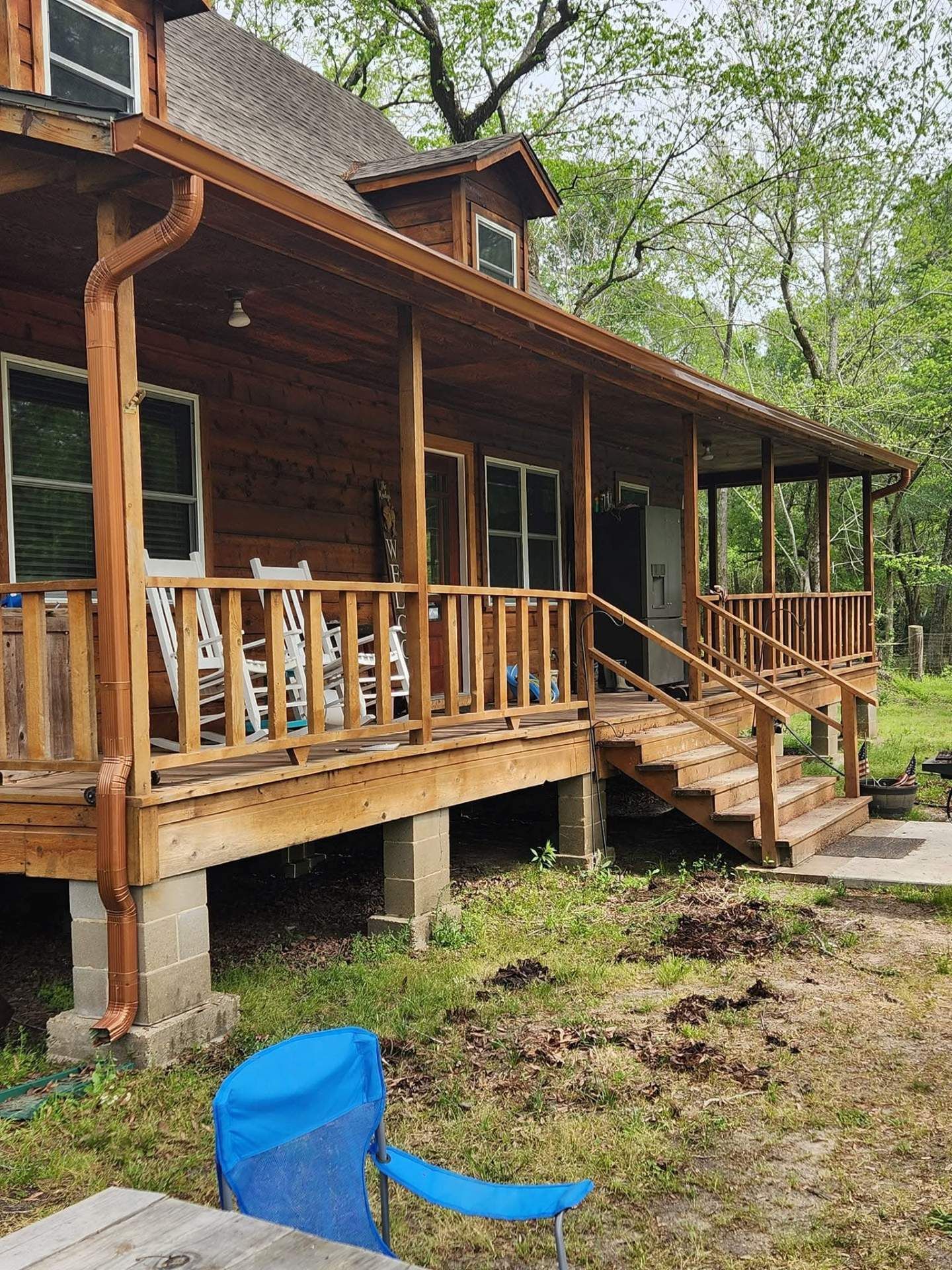 Wooden cabin with porch, railing, and stairs. Blue chair sits in the yard.