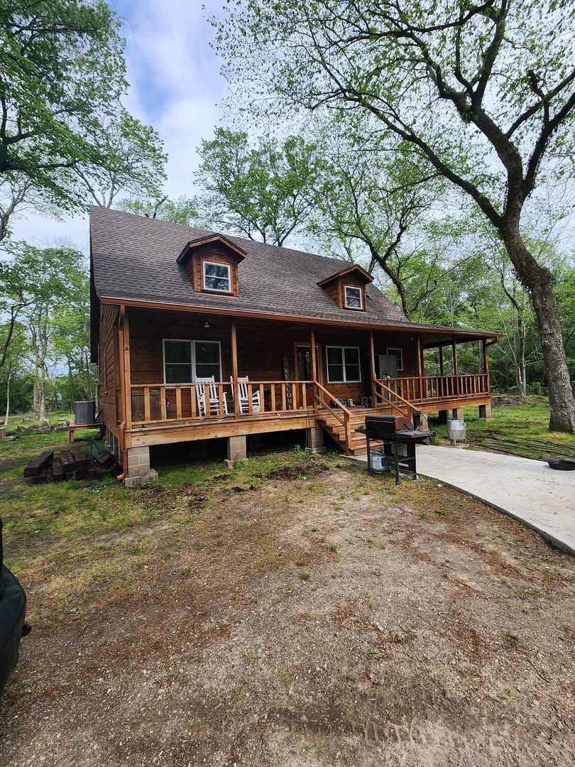 Log cabin with porch and dormer windows, surrounded by trees and a concrete path.