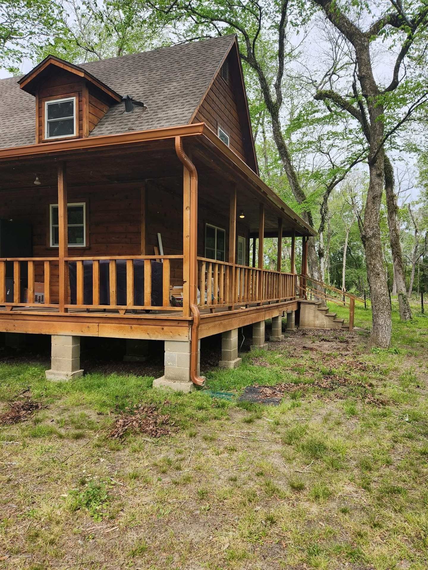 Wooden cabin with porch on concrete pillars; surrounded by trees and grass.