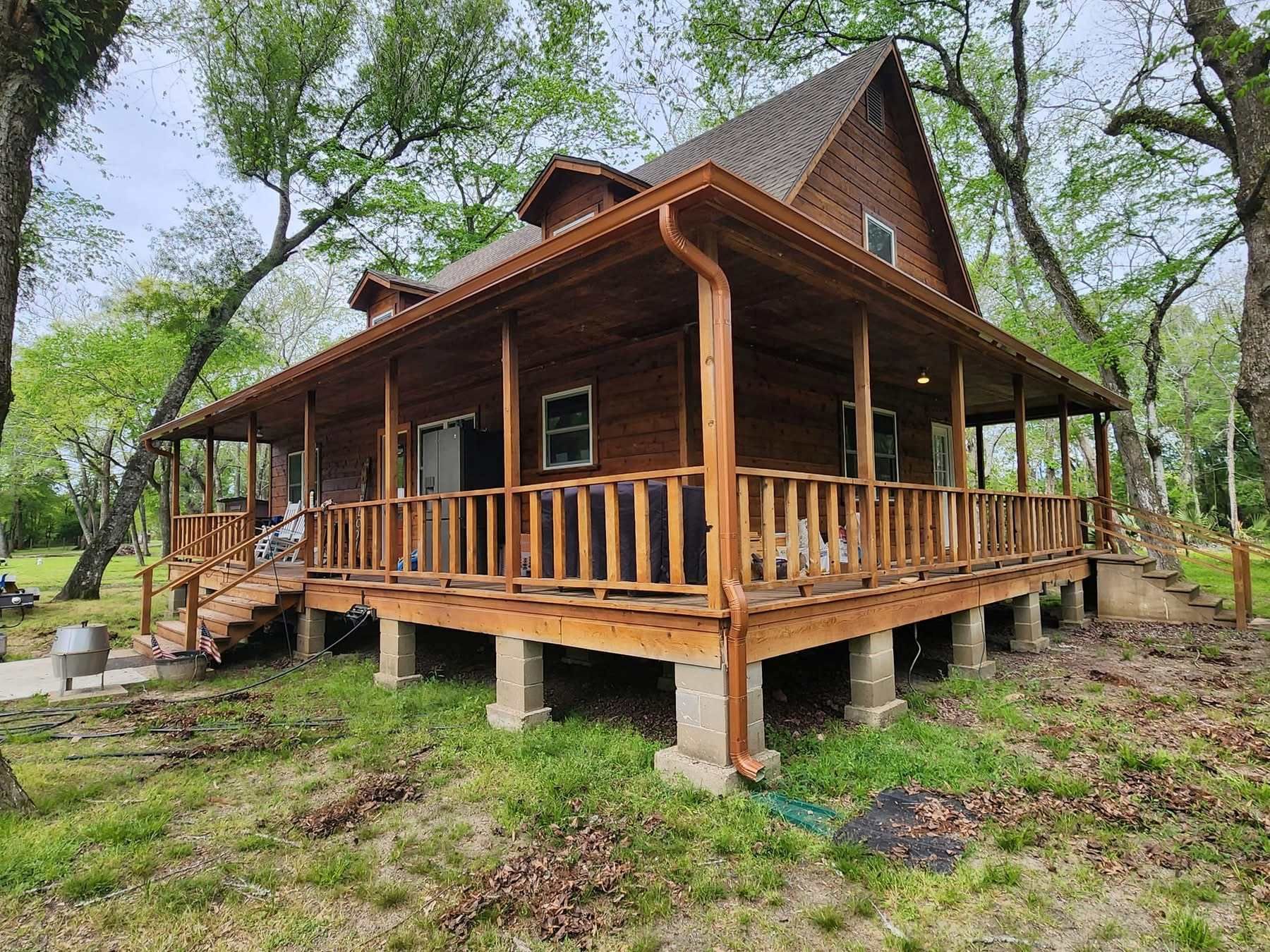 Wooden cabin with a wrap-around porch, on stilts, surrounded by trees and greenery.