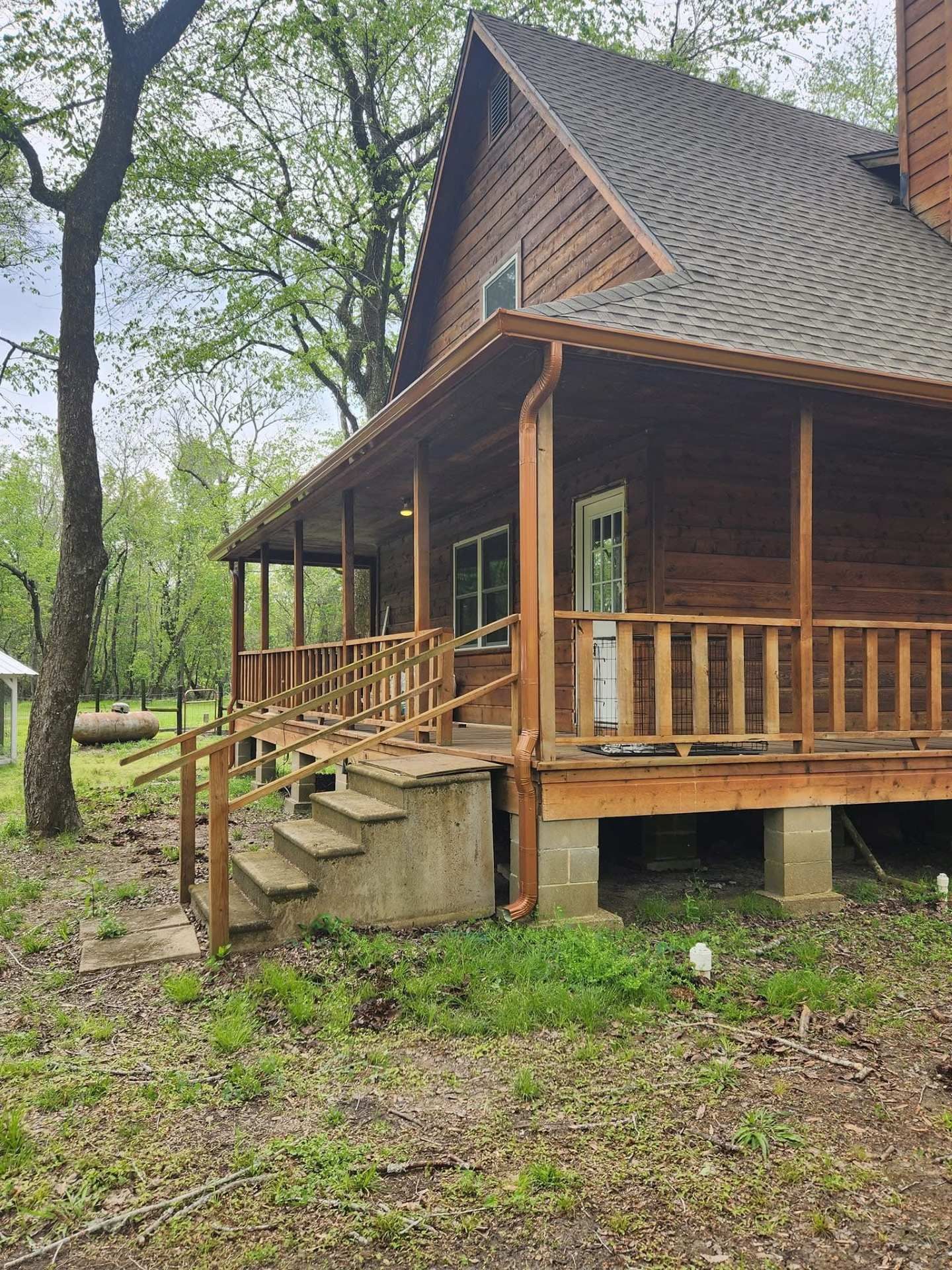 Wood-sided house with covered porch, wooden railing, and concrete steps in a wooded setting.