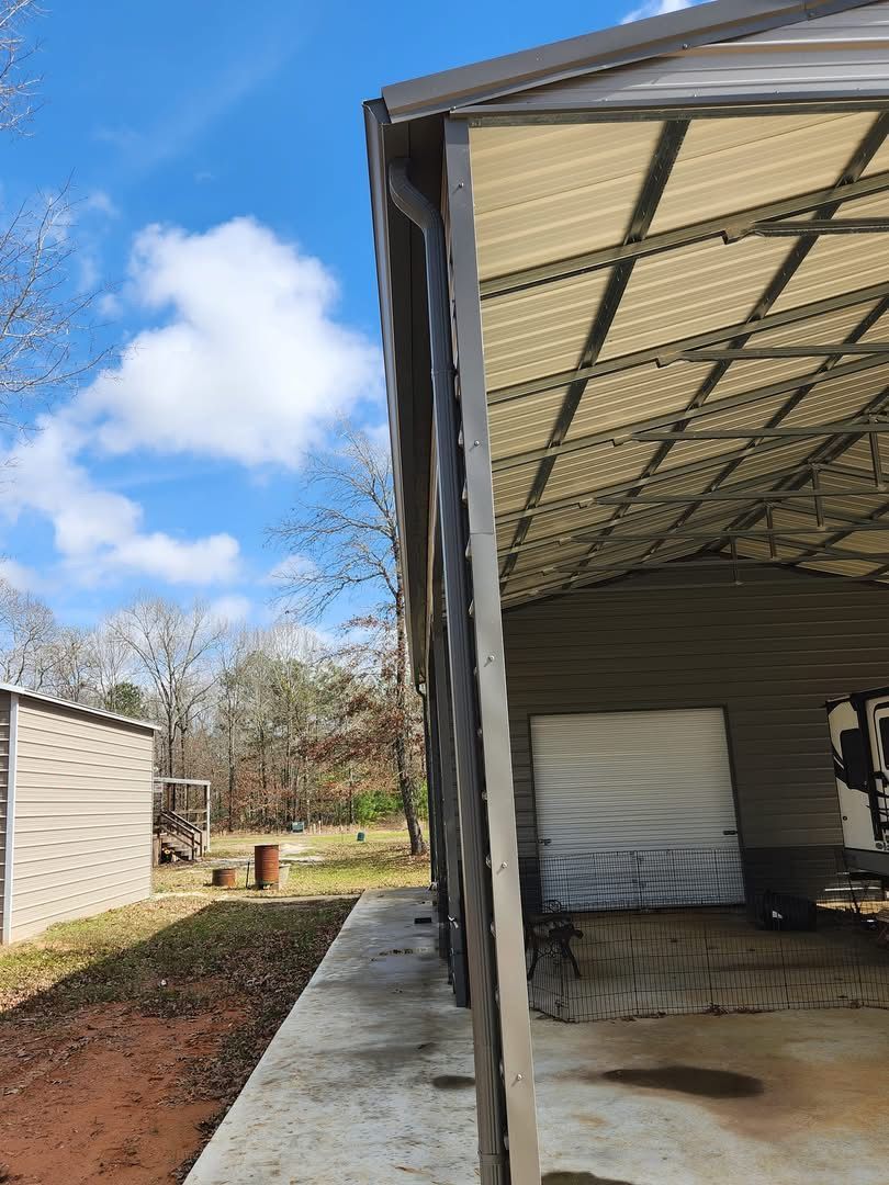 Metal carport with beige roof, concrete floor, and attached building in a grassy outdoor setting with blue sky.