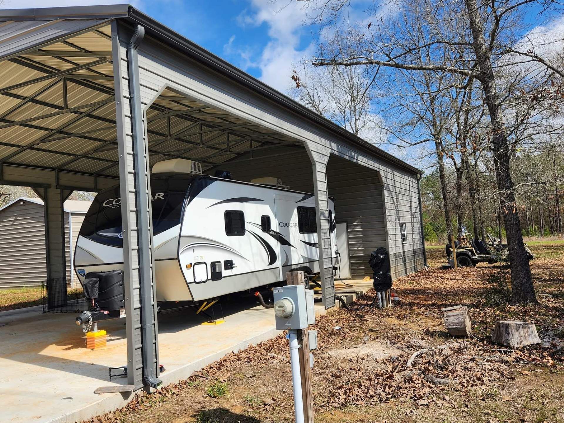 RV under gray metal carport in wooded area. Electrical hookup visible.