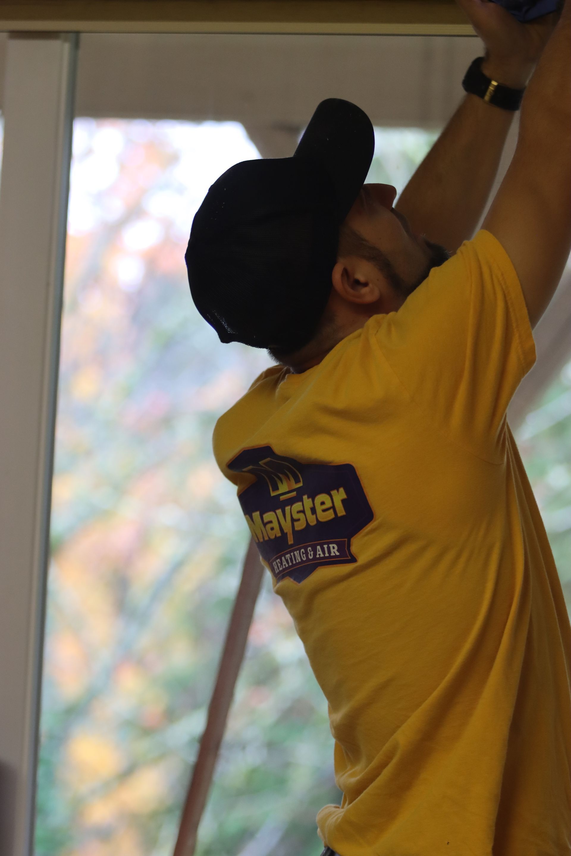 Person wearing a yellow shirt and black cap reaching up to work on a window.