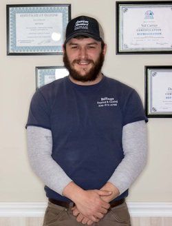 A man with a beard wearing a baseball cap and a blue shirt with a company logo, posing in front of framed certificates.