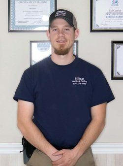 Man in a dark blue shirt and baseball cap, standing in front of certificates. He has a goatee and is smiling.