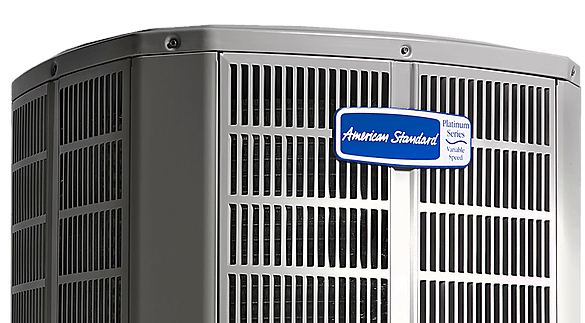 Close-up of an American Standard air conditioning unit with a blue and white brand tag in the center. The unit is gray and has a slatted design.