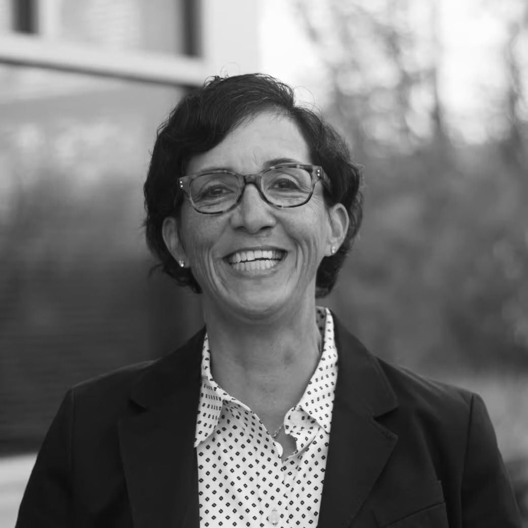 Woman with glasses, smiling, in a blazer and patterned shirt, outdoors.