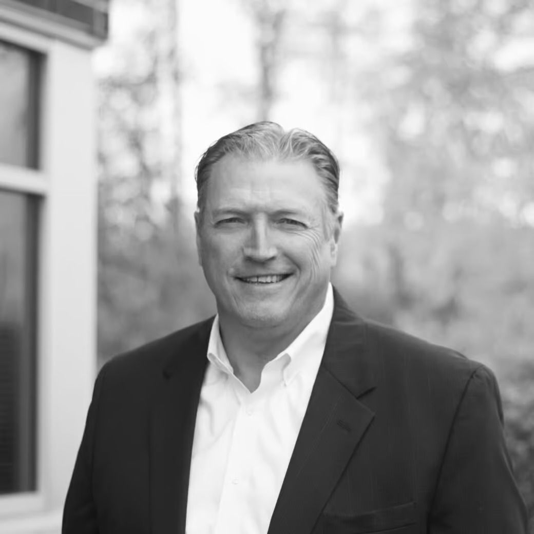 Man in suit jacket smiles, standing outdoors near a building with a window.