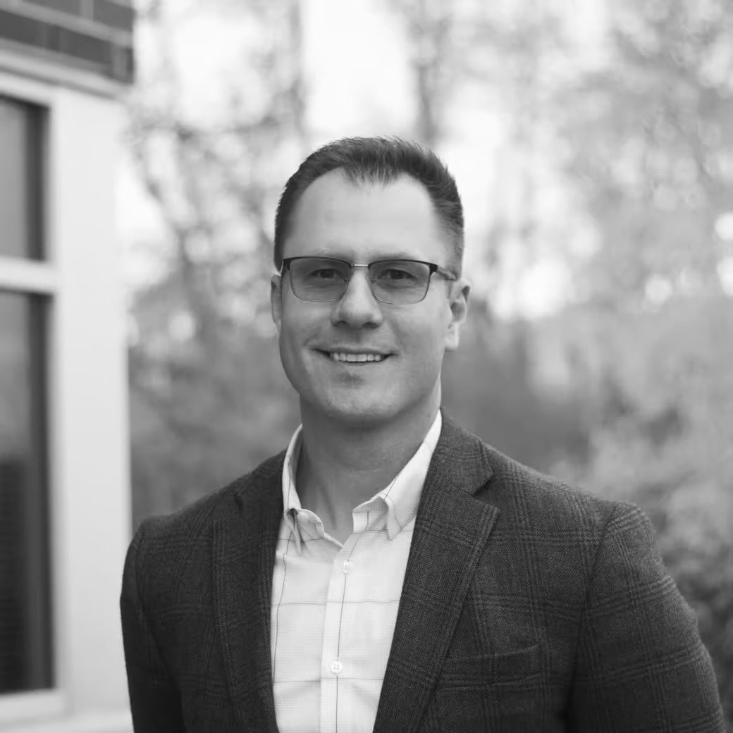 Man in blazer and glasses smiles outdoors.