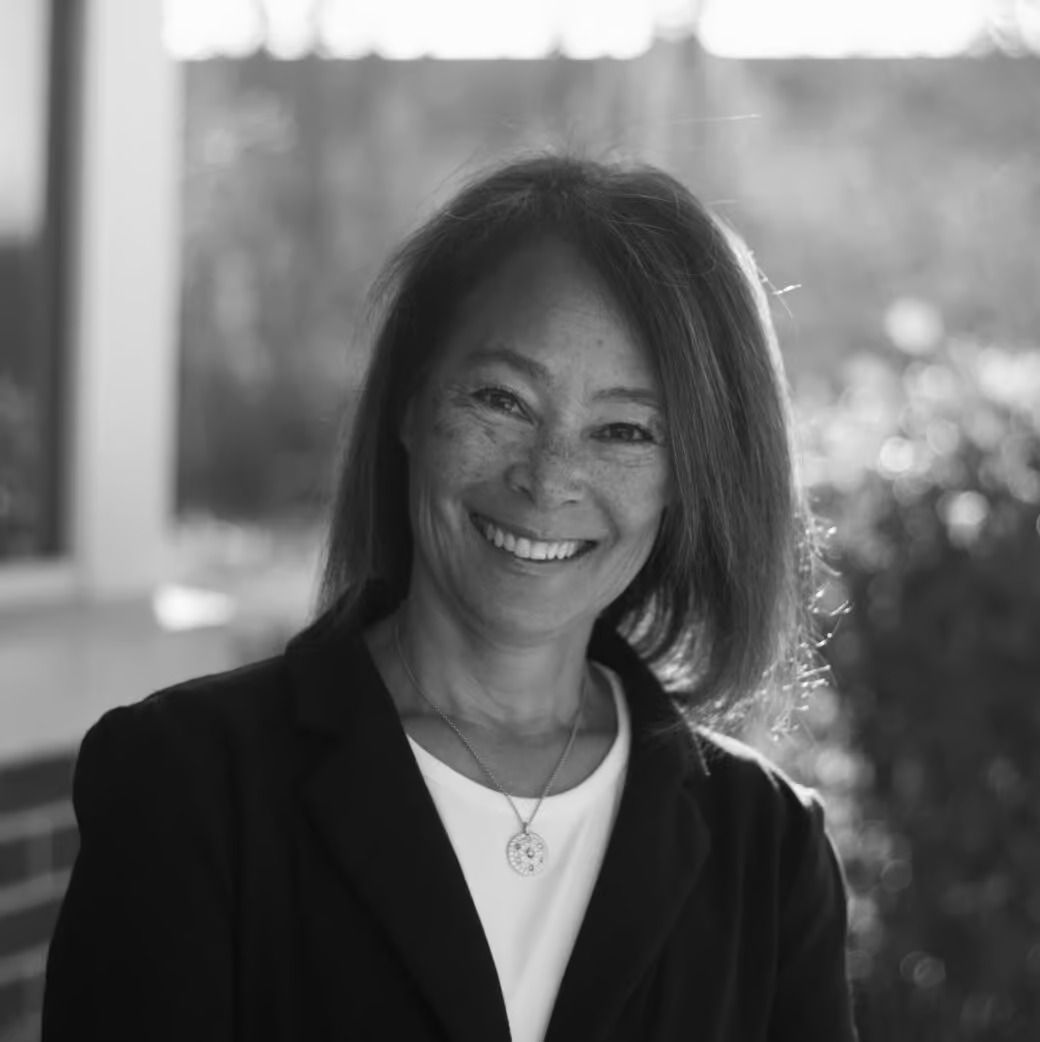 Smiling woman, black and white, wearing a blazer, white shirt, and necklace; outdoors with blurred background.