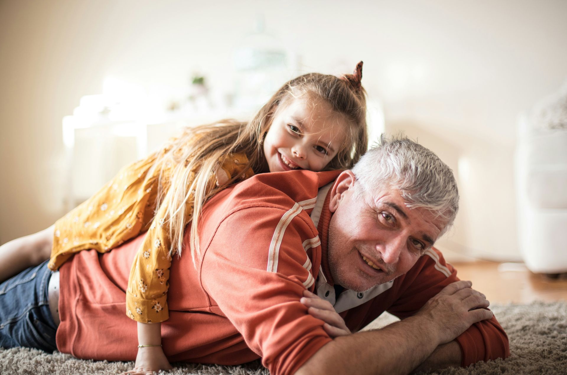 Girl rides on a person's back while they both lay on a rug, smiling.