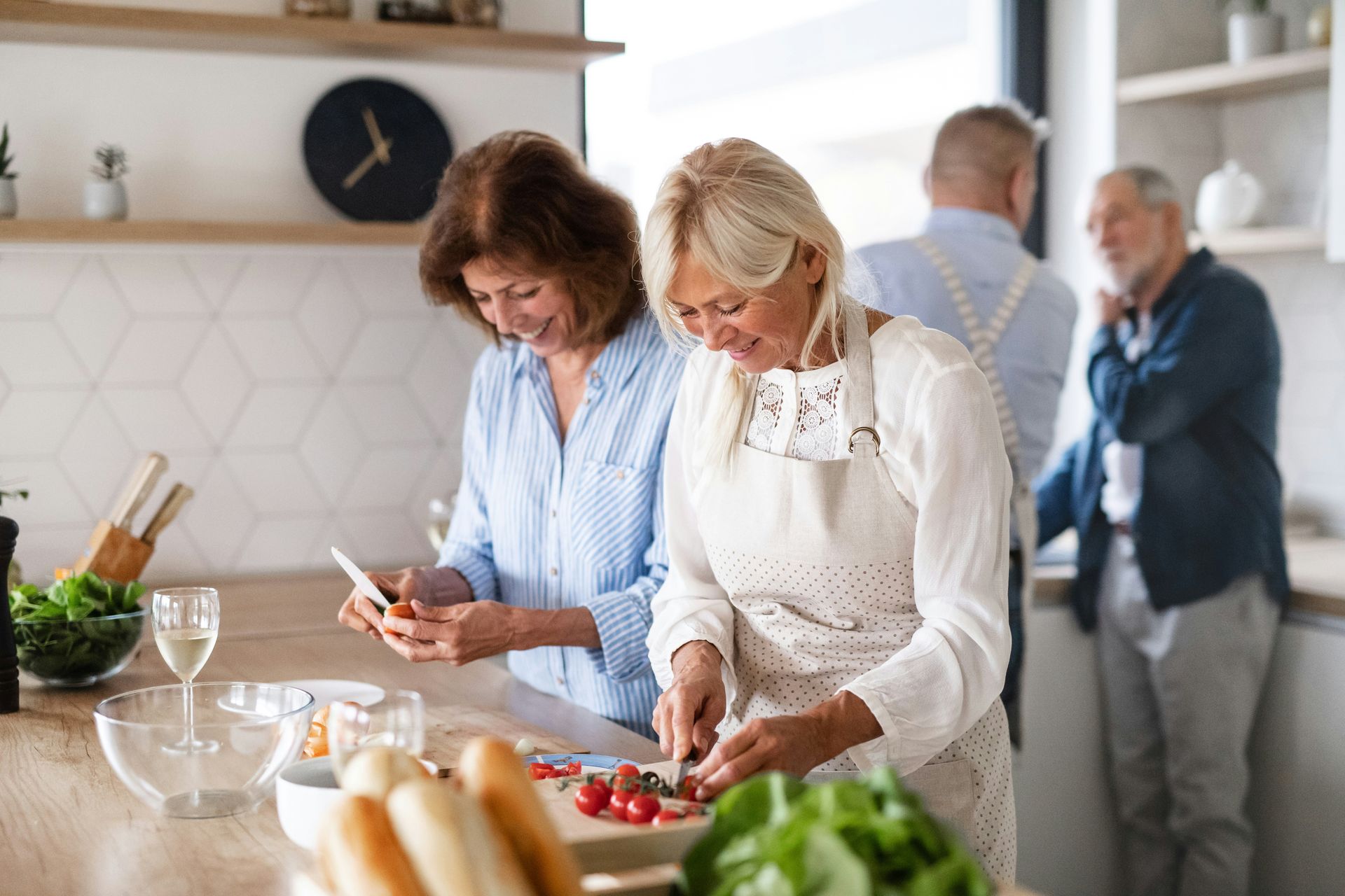 Two women cook in a kitchen with two men nearby. One woman slices vegetables, the other looks at a phone.