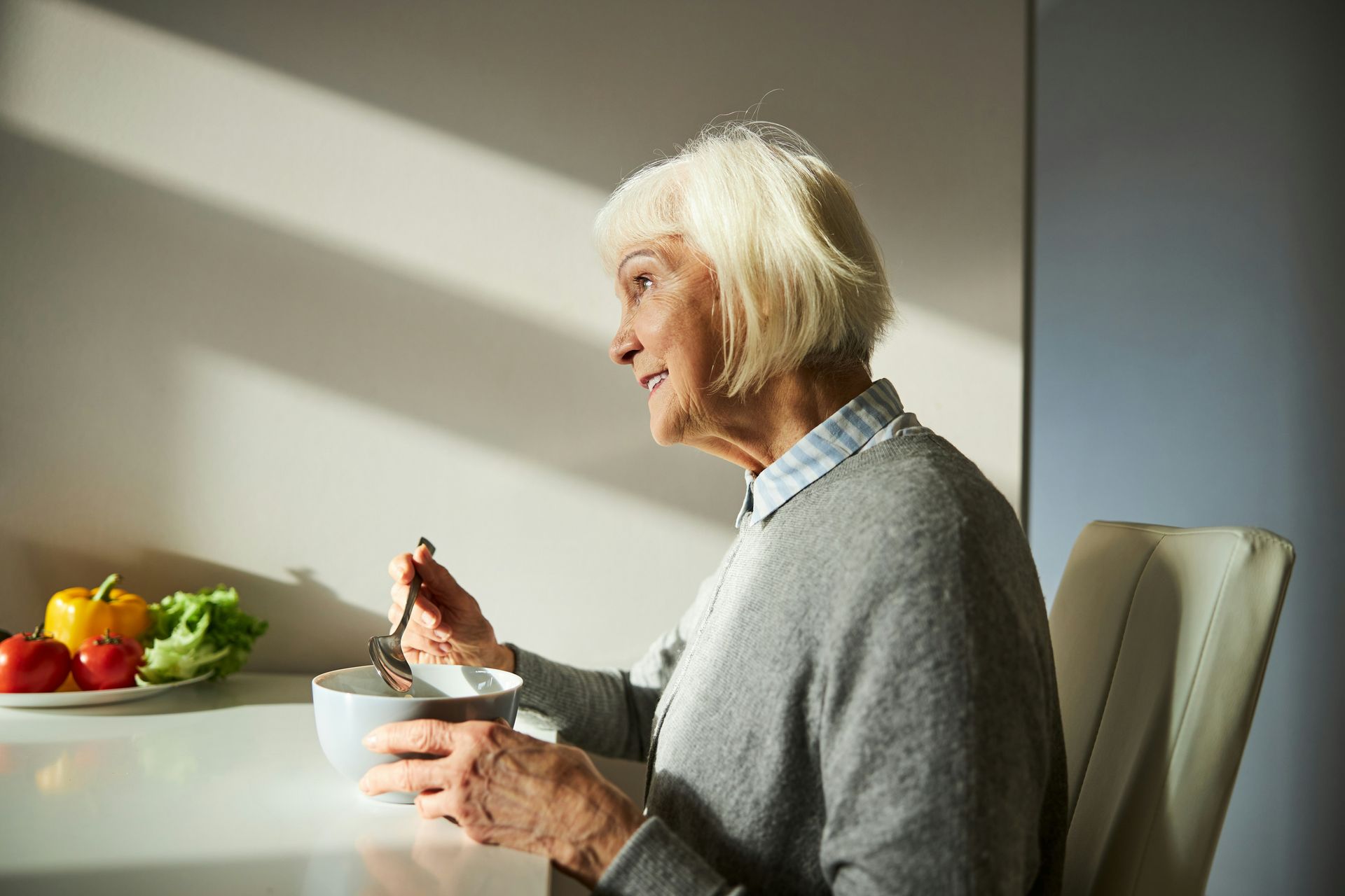 Elderly person seated at table eating from a bowl. Side profile, smiling, sunlight, vegetables nearby.