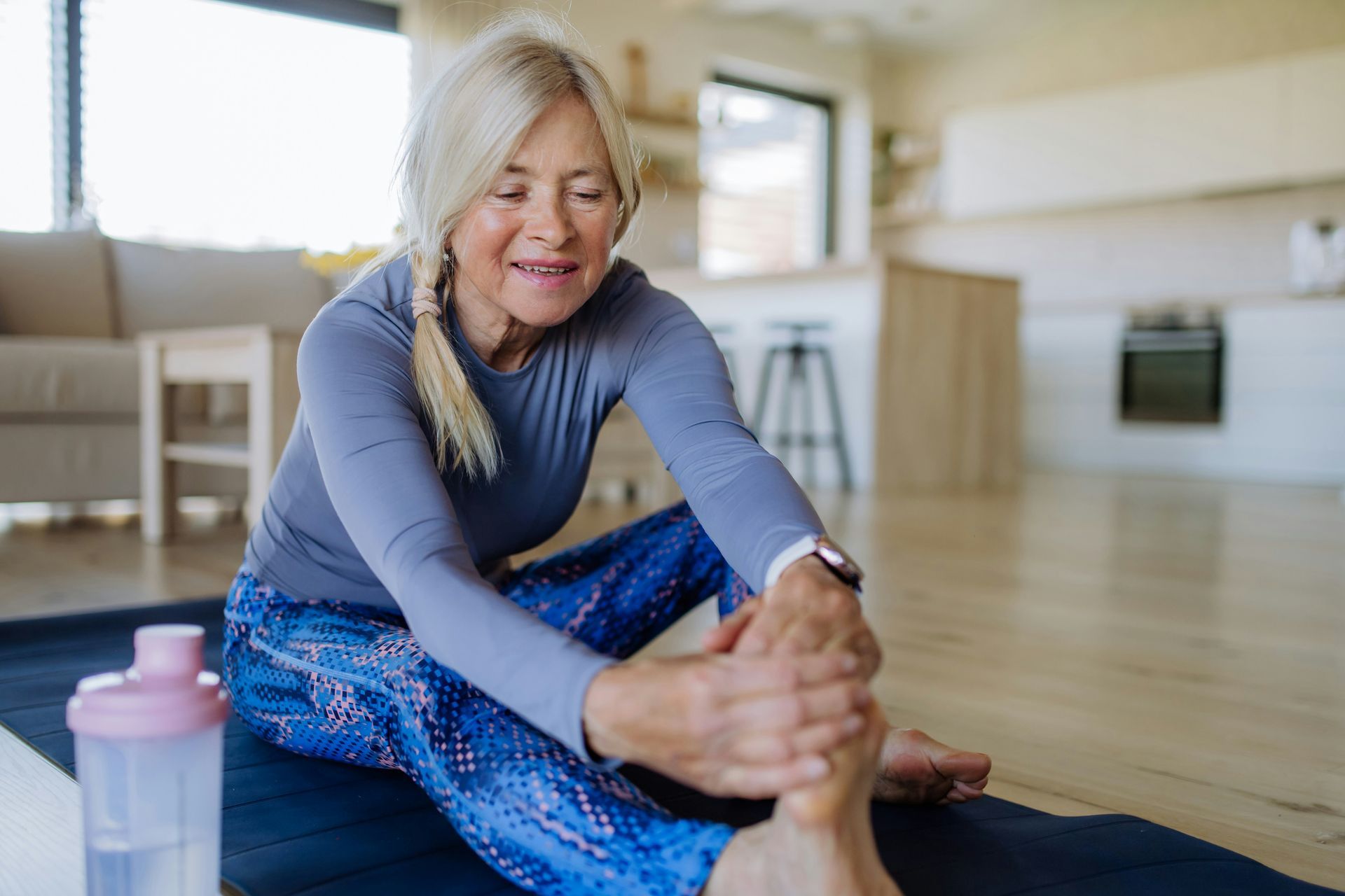 Woman stretching on yoga mat, indoors; wearing blue athletic wear. She's holding her foot, looking down.