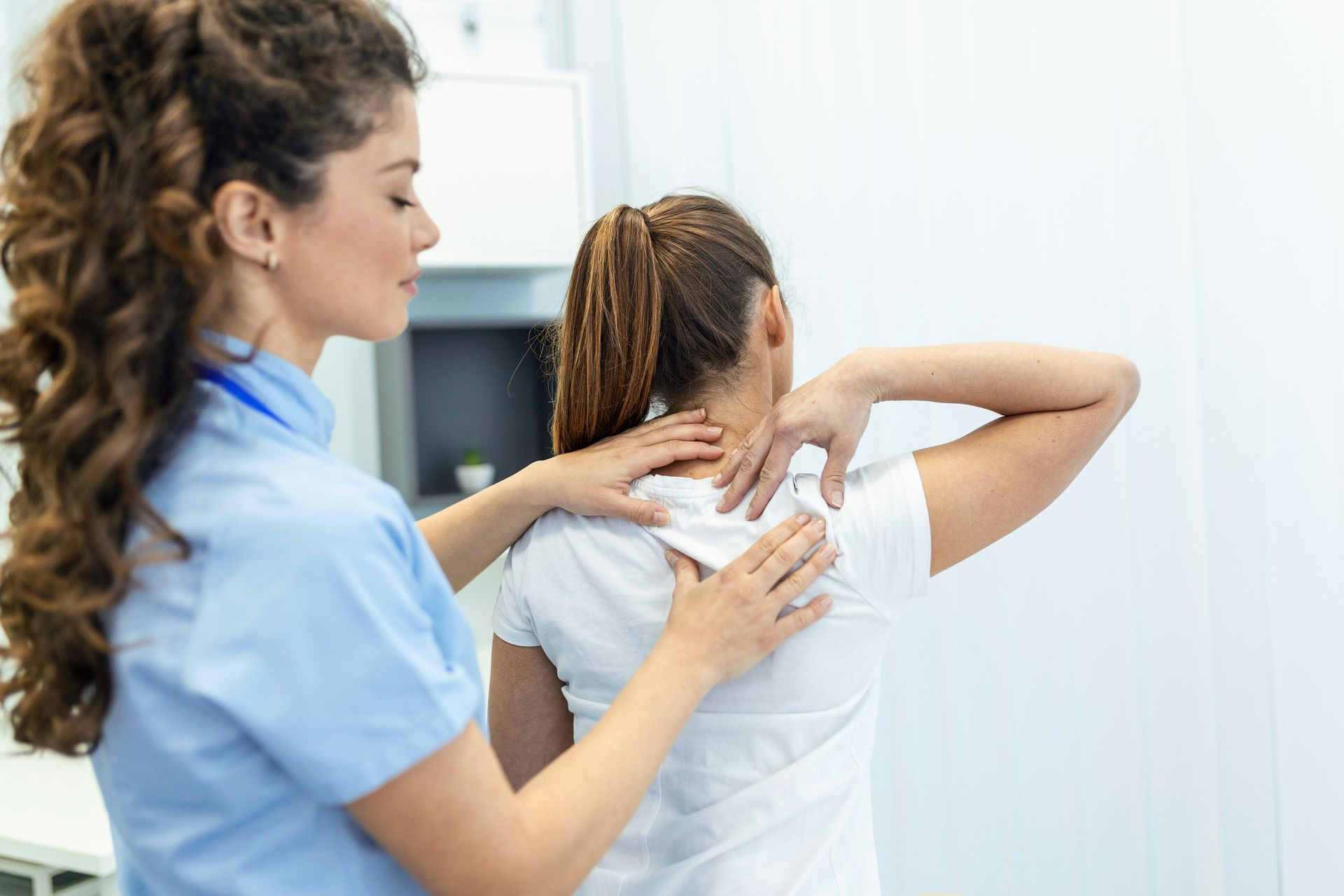 A medical professional examines a patient's back. The patient wears a white shirt in a doctor's office.